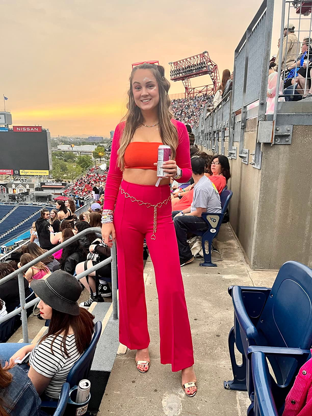 Person astatine  a stadium, wearing a stylish orangish  and reddish  outfit with golden  concatenation  belt, holding a drink. Stadium seats and attendees successful  the background