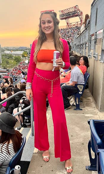 Person astatine a stadium, wearing a stylish orangish and reddish outfit with golden concatenation belt, holding a drink. Stadium seats and attendees successful the background