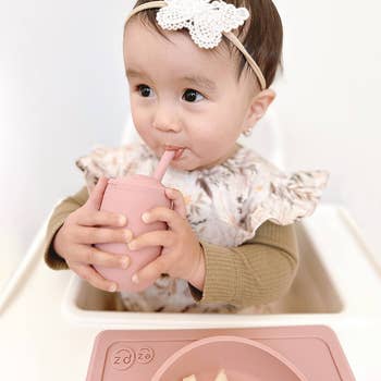 Toddler with headband using a sippy cup, wearing a ruffled outfit, at a mealtime setting