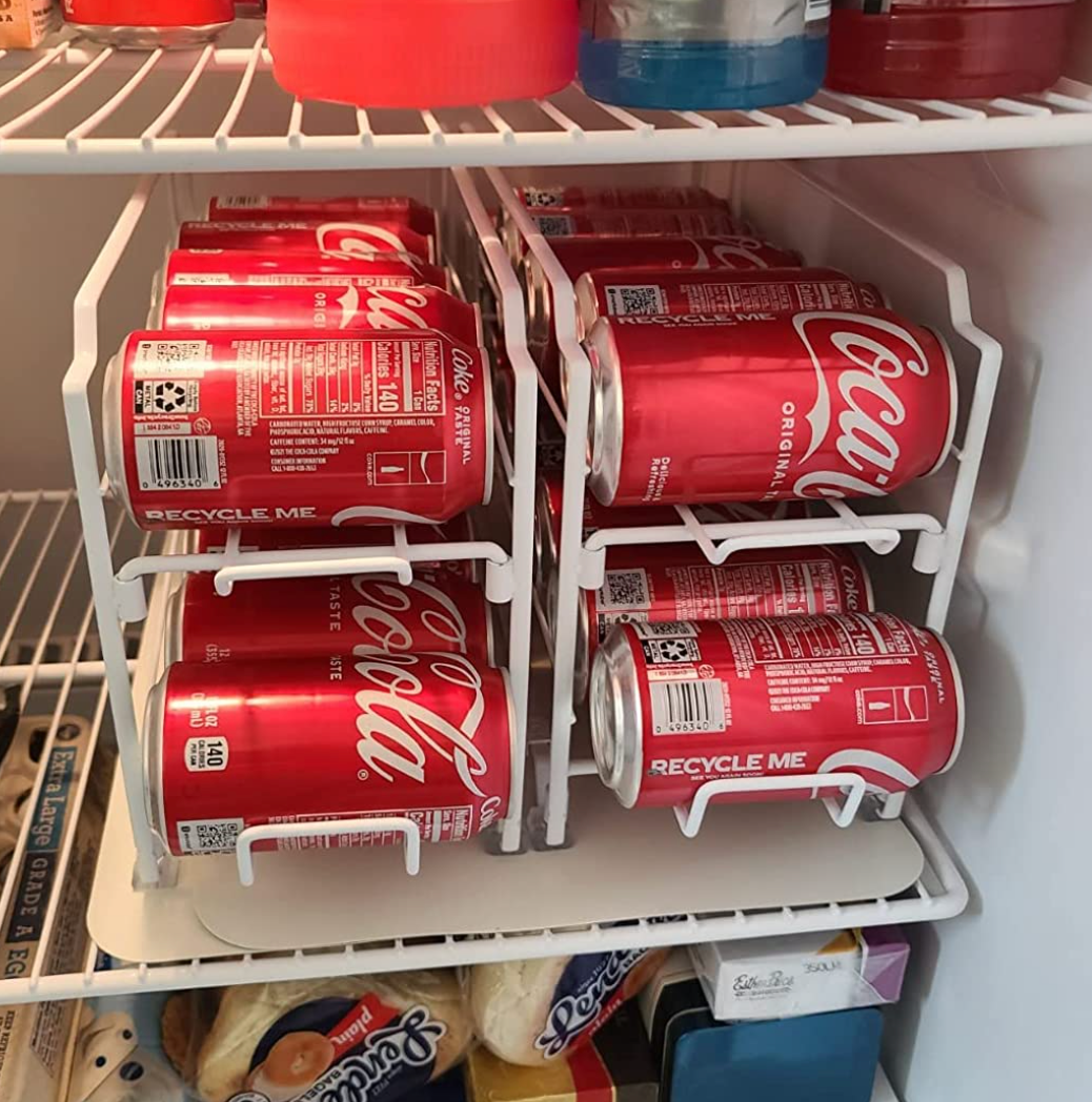 A set of coke cans stored horizontally in the two white wire frame dispensers on a fridge shelf 