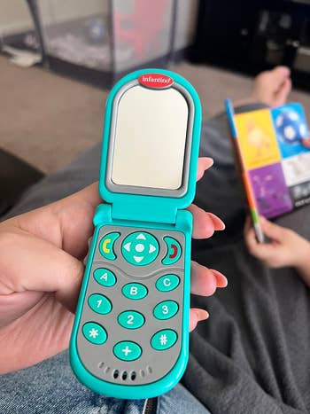 Toy flip phone held by a person, resembling a real keypad and screen. Background shows blurred scene with a person reading. Ideal for children's playtime