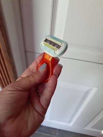 Hand holding an orange razor with a multi-blade head in front of a white door