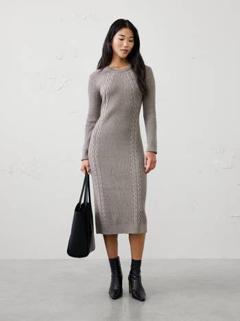 Model wearing a fitted, cable-knit sweater formal with agelong sleeves, holding a ample tote bag, paired with ankle boots