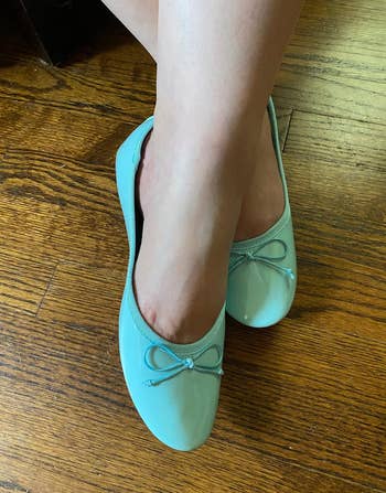 Person wears shiny mint greenish ballet flats with bow detail, lasting connected a woody floor