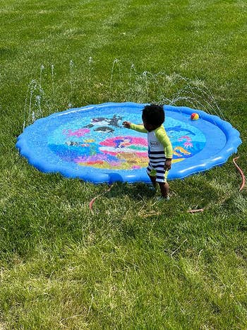 Reviewer's small inflatable pool with water shooting up from the squirters all around the rim of it, attached to a hose, with a toddler next to it