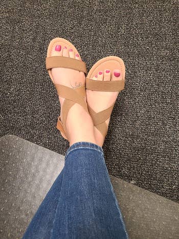 Reviewer wearing strappy sandals with pinkish  toenail polish rests her feet connected  a carpeted floor, showcasing casual footwear style