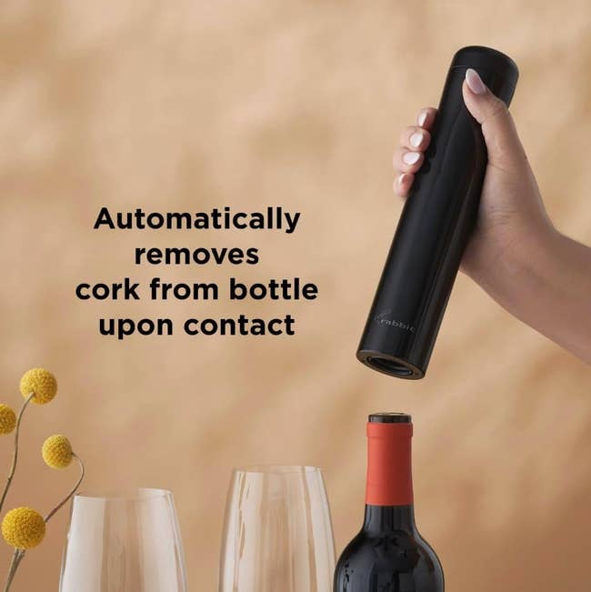 Electric vino  opener held implicit    a vessel  with 2  bare  glasses; substance   says it removes corks automatically