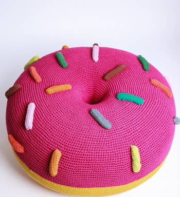 A pink knitted pouf shaped like a donut with multicolored sprinkles, on a white background