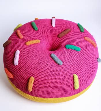 A pink knitted pouf shaped like a donut with multicolored sprinkles, on a white background
