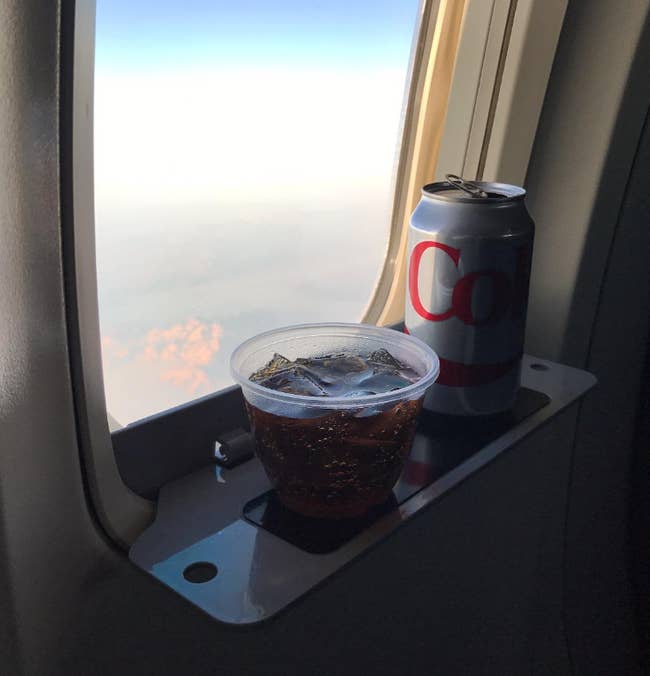 reviewer image of plane window with shelf attached. plastic cup and can of coke on shelf. 