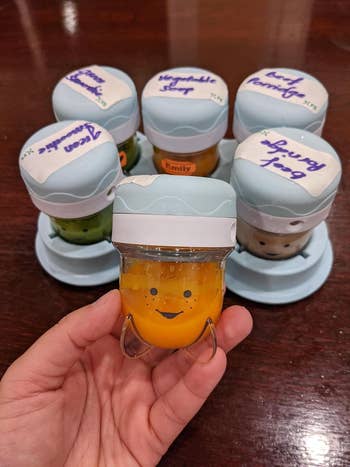 Tangan memegang toples makanan bayi dengan wajah tersenyum, dikelilingi oleh lima stoples berlabel dengan rasa makanan bayi yang berbeda