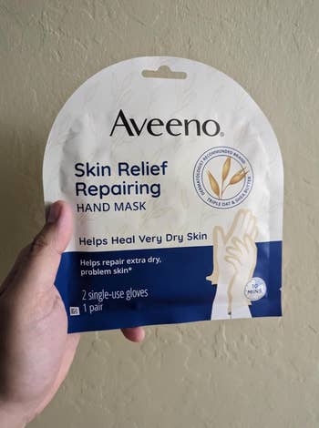 Person holding Aveeno Skin Relief Repairing Hand Mask packaging for precise  adust  skin. Advertises 2  single-use gloves and 10-minute use