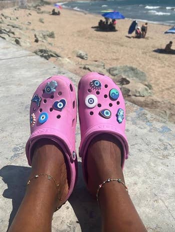 Close-up of two feet wearing pink Crocs adorned with various charms, with a beach, ocean, and sunbathers blurred in the background