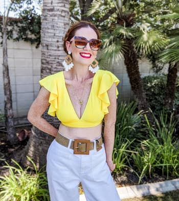 Reviewer wearing ruffled yellow top, white pants, large belt, sunglasses, and statement earrings