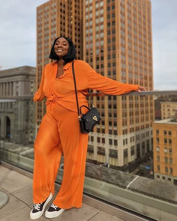 reviewer in a stylish orange jumpsuit with a shoulder bag, posing confidently in an urban setting