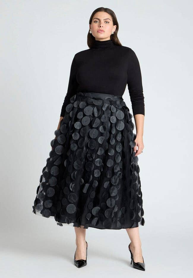 Person wearing a stylish black turtleneck and a polka-dot textured skirt with black heels, posing confidently for a fashion article