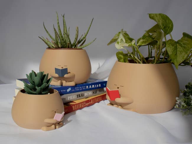 Three ceramic pots shaped similar  seated figures, each   holding a book, with plants inside. Books stacked nearby. Ideal for publication  and works  lovers
