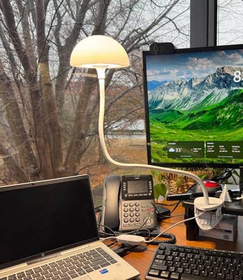 Desk with a laptop, phone, keyboard, and monitor showing a mountain landscape; a modern mushroom-shaped lamp is attached to the desk