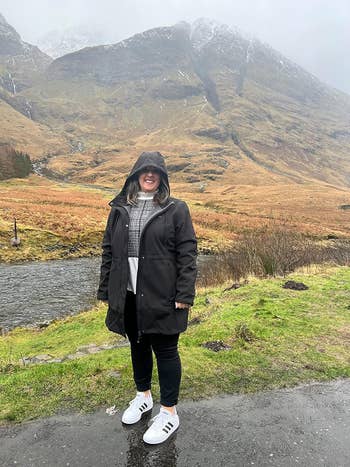 Person in a raincoat and sneakers standing in front of a mountain landscape