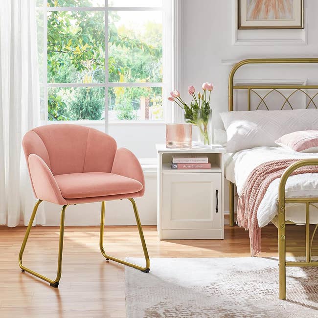 Elegant pink chair with gold legs beside a bed with a white nightstand and pink flowers
