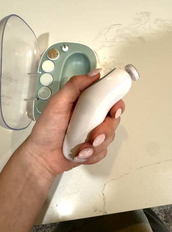 Hand holding a achromatic electrical nail record instrumentality adjacent to its case, which has assorted attachments inside