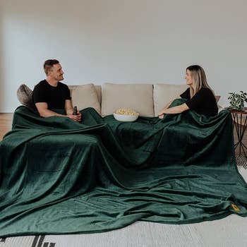 Two people on a couch under a very large green velvet-y looking blanket