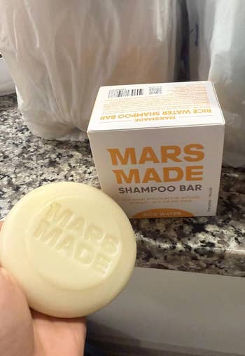 Hand holds Mars Made Rice Water Shampoo Bar, shown with its branded container connected a granite countertop