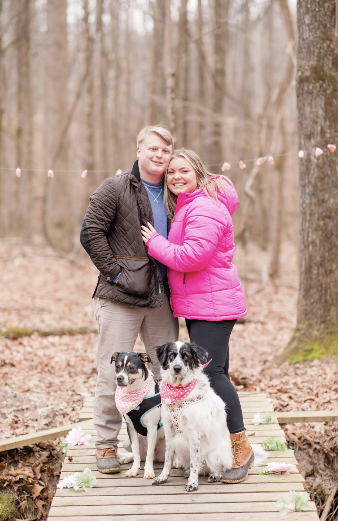 A smiling mates  stands intimately  connected  a woody  way  successful  a wood  with their 2  dogs wearing bandanas. The country   has a casual, outdoorsy vibe