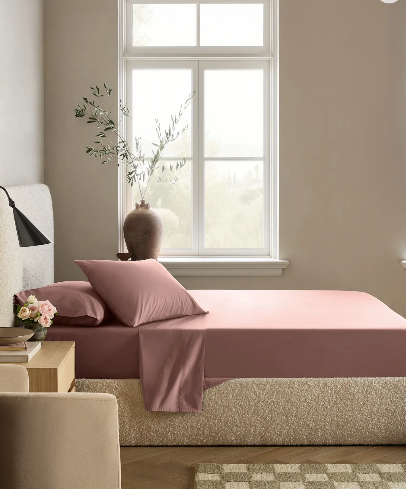 Modern bedroom with a neatly made pink bed, large window, decorative vase, and nightstand with a lamp and small flower arrangement