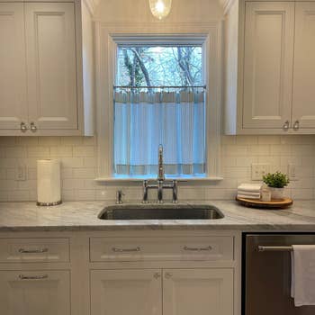 Modern kitchen with white cabinets, marble countertop, and under-mount sink. A small window above the sink with a pleated shade. Decor includes a small plant and towels