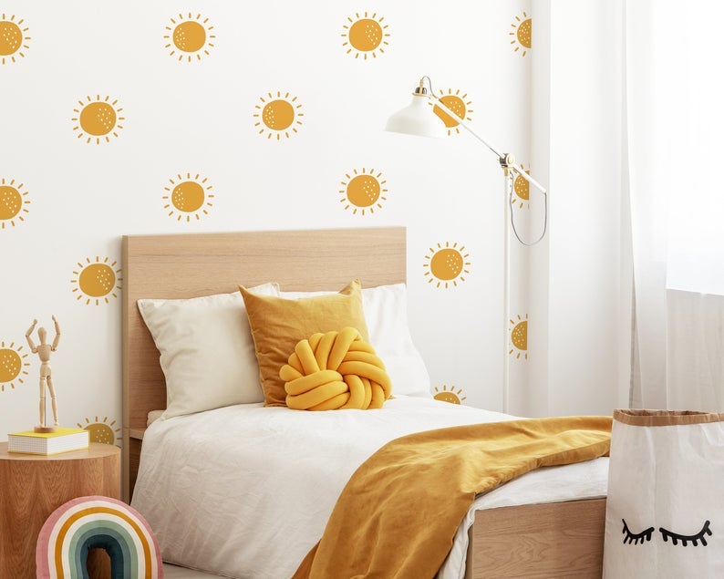 a bed with a wooden frame, gold yellow pillows, a yellow throw across the end, and a white blanket on top with a white wall behind it and the sunshine decals all over the wall behind it kind of like large polka dots