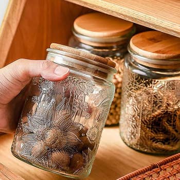 Hand holding an intricately designed glass jar with a wooden lid in front of two similar jars on a wooden shelf
