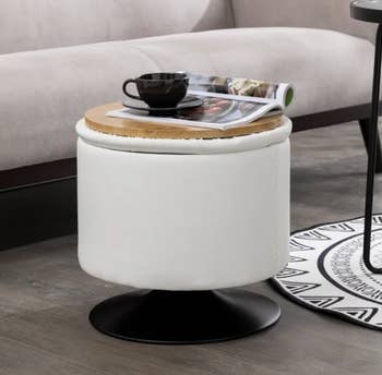 The storage ottoman in white with the lid flipped so the wooden try side is up with a magazine and coffee mug sitting on it