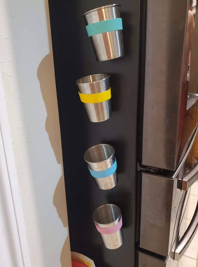 A set of four cups magnetized to the side of the fridge with different color bands around them 