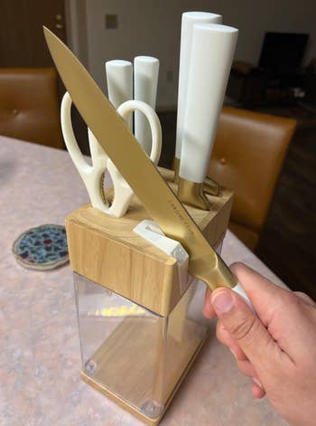 A hand holds a gold-tone kitchen knife in front of a wooden knife block set on a countertop, featuring multiple knives and a pair of scissors