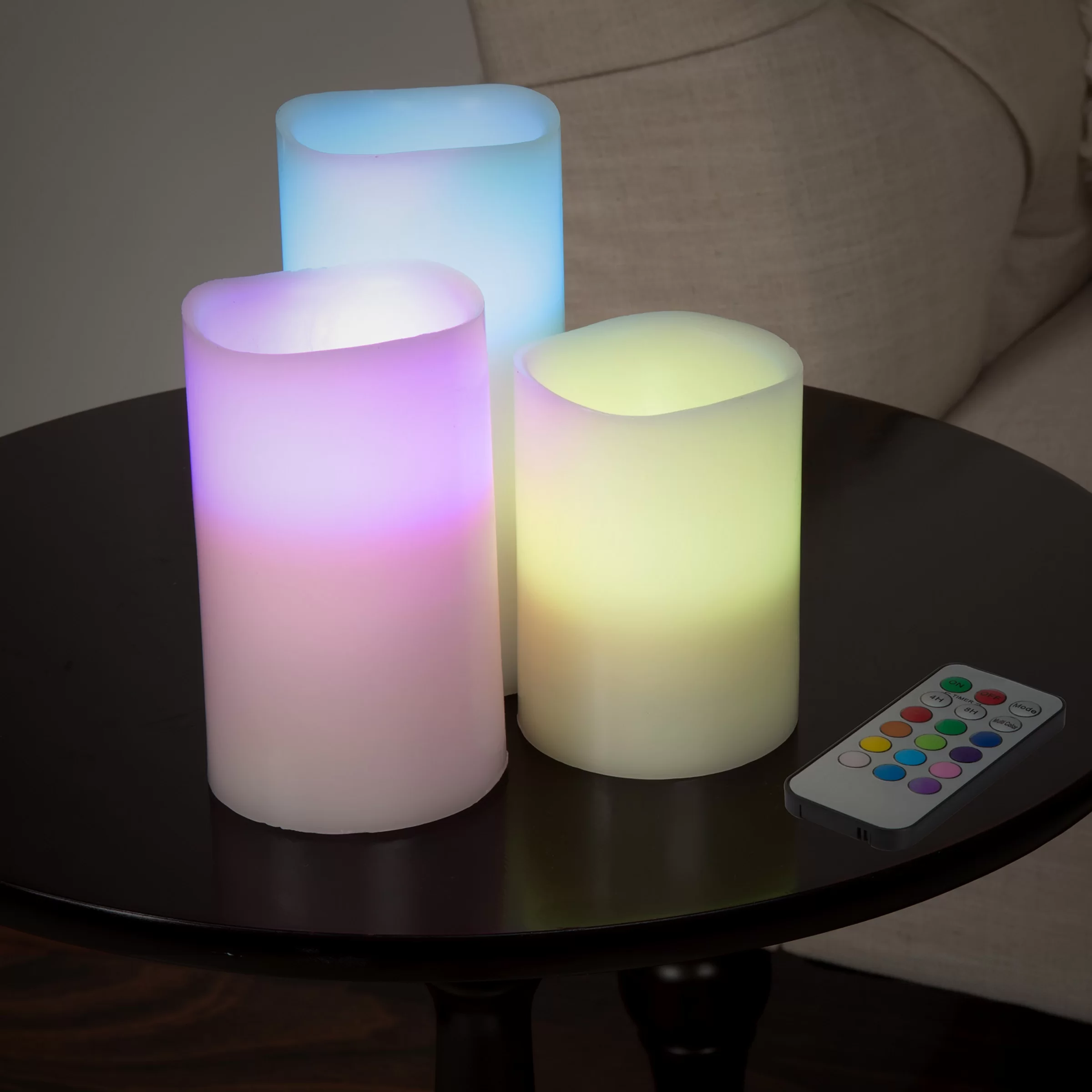 Three color-changing flameless candles in purple, yellow, and blue on top of black side table next to small silver remote