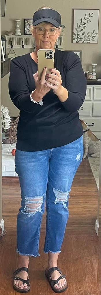 A reviewer taking a mirror selfie wearing a black long-sleeve top, blue ripped jeans, black sandals, and a black cap.