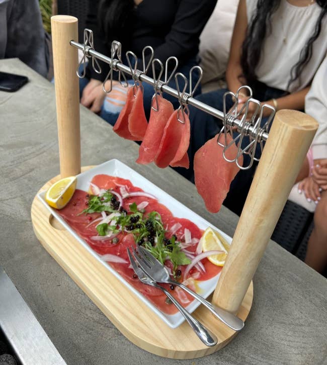 Sliced cured meats suspended from clips on a stand beside garnished meat on a plate, with people seated in the background