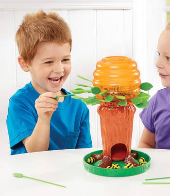 Two young children, a boy in a blue shirt and a girl in a purple shirt, are playing with a tree-themed game, placing leaves into slots