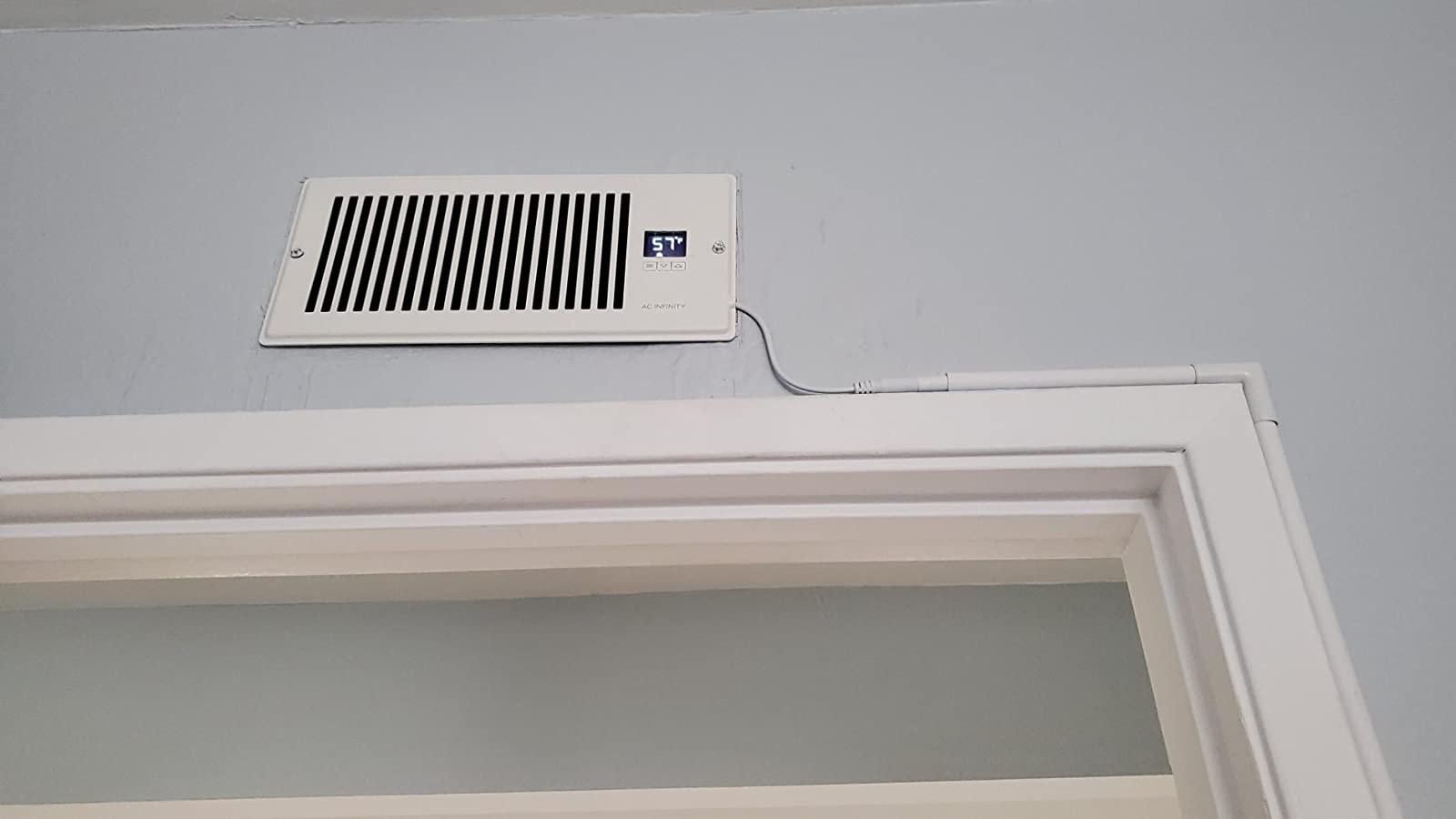 a reviewer photo of the booster fan installed on a wall vent with the cord hugging the doorway molding 