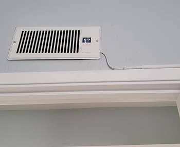 a reviewer photo of the booster fan installed on a wall vent with the cord hugging the doorway molding 
