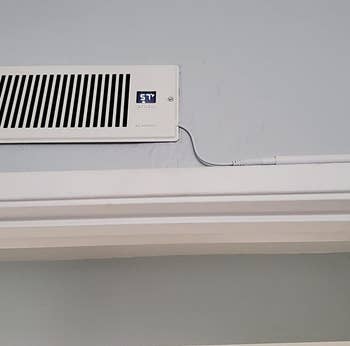 a reviewer photo of the booster fan installed on a wall vent with the cord hugging the doorway molding 