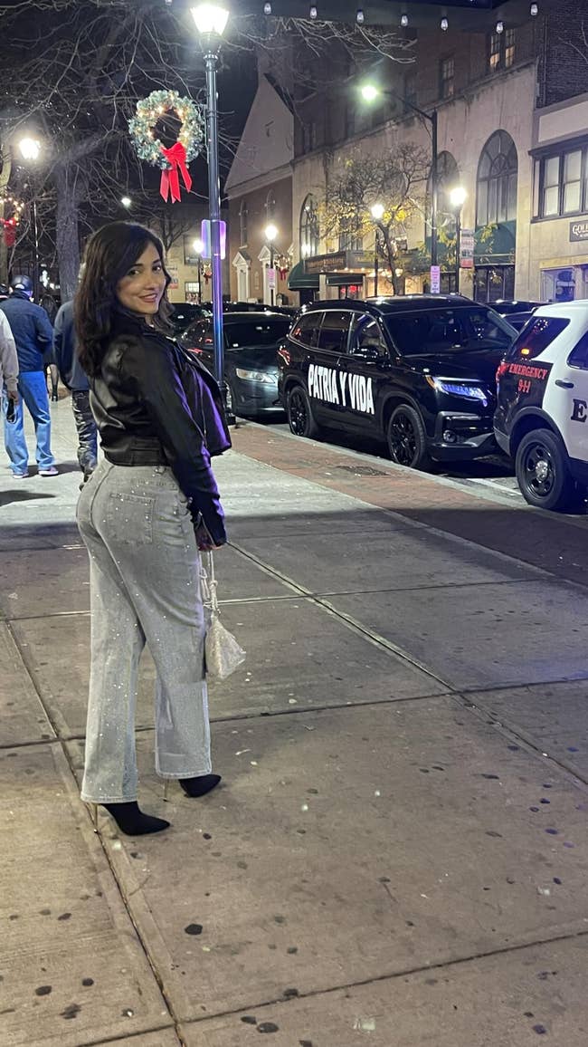 A idiosyncratic poses connected a metropolis sidewalk astatine night, wearing a stylish leather overgarment and sparkling pants, with parked constabulary cars successful the background