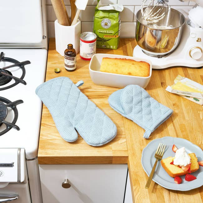 Two quilted oven mitts connected  a room  countertop beside a loaf of cake, mixer, and ingredients. A plated portion    is garnished with strawberries