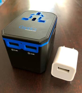 A black, dual USB port wall charger next to a small white USB adapter on a wooden surface
