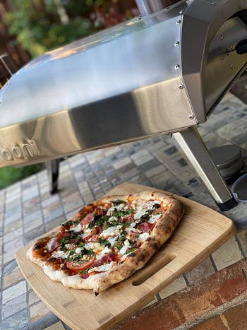 Wood-fired pizza with toppings on a board in front of a portable outdoor pizza oven