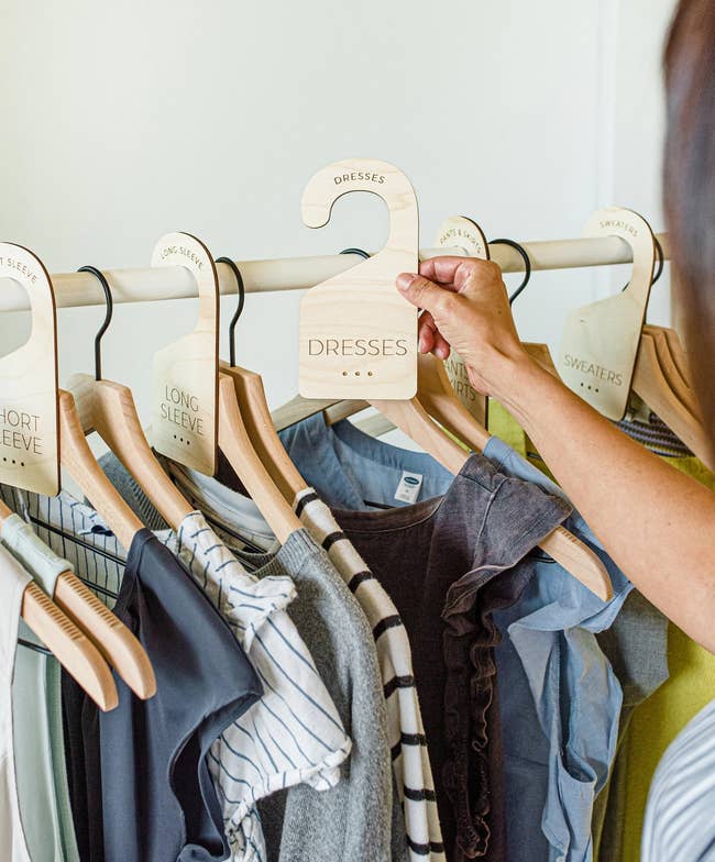 person holding wooden closet divider labeled 'dresses' above hanging clothes