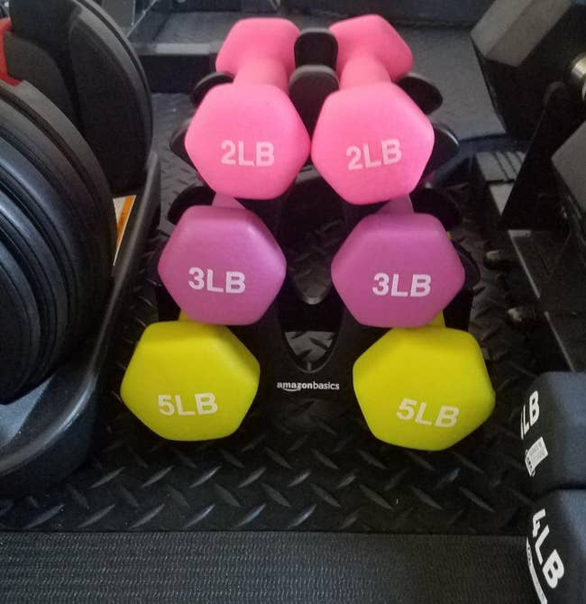 reviewer pic of set of pink 2-pound weights, dark pink 3-pound weights, and yellow 5-pound weights on black stand