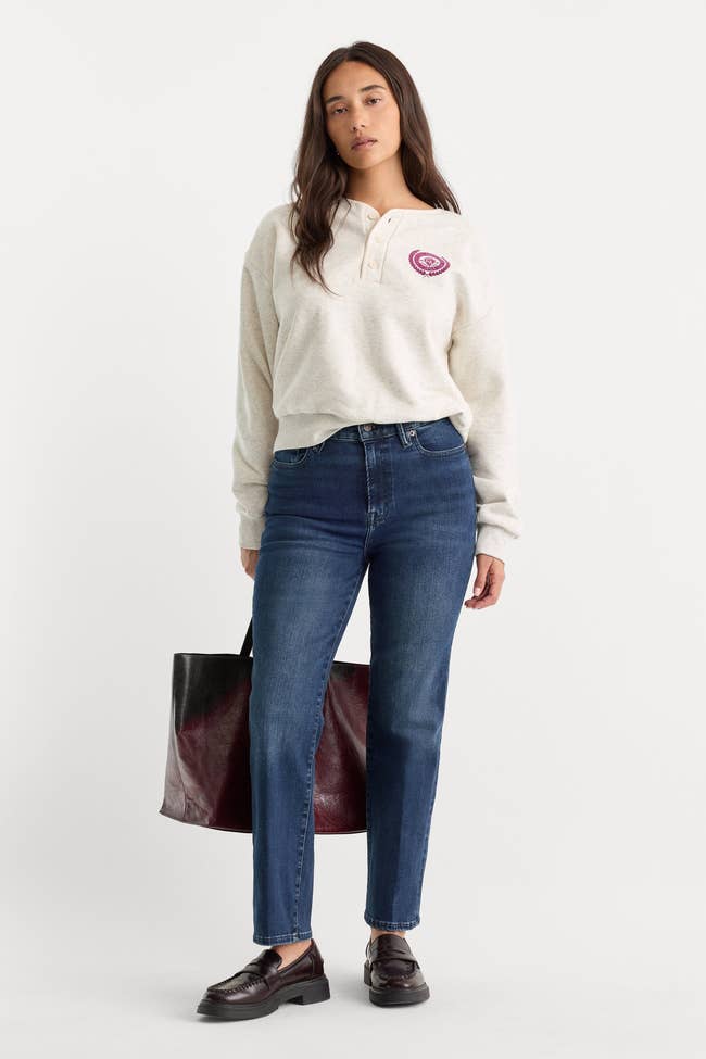  pick  henley garment  with emblem, bluish  jeans, brownish  loafers, holding ample  acheronian  tote
