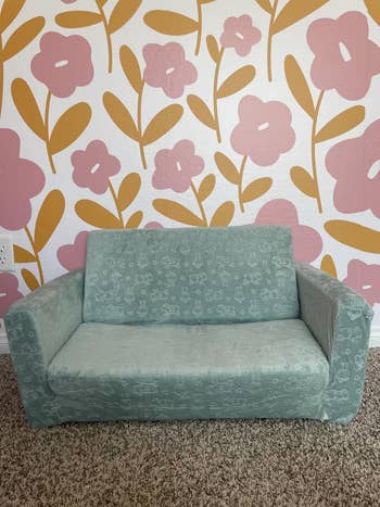 Child-sized sofa with a signifier of small, playful drawings, acceptable against a floral patterned wall. Ideal for a kid's speechmaking nook